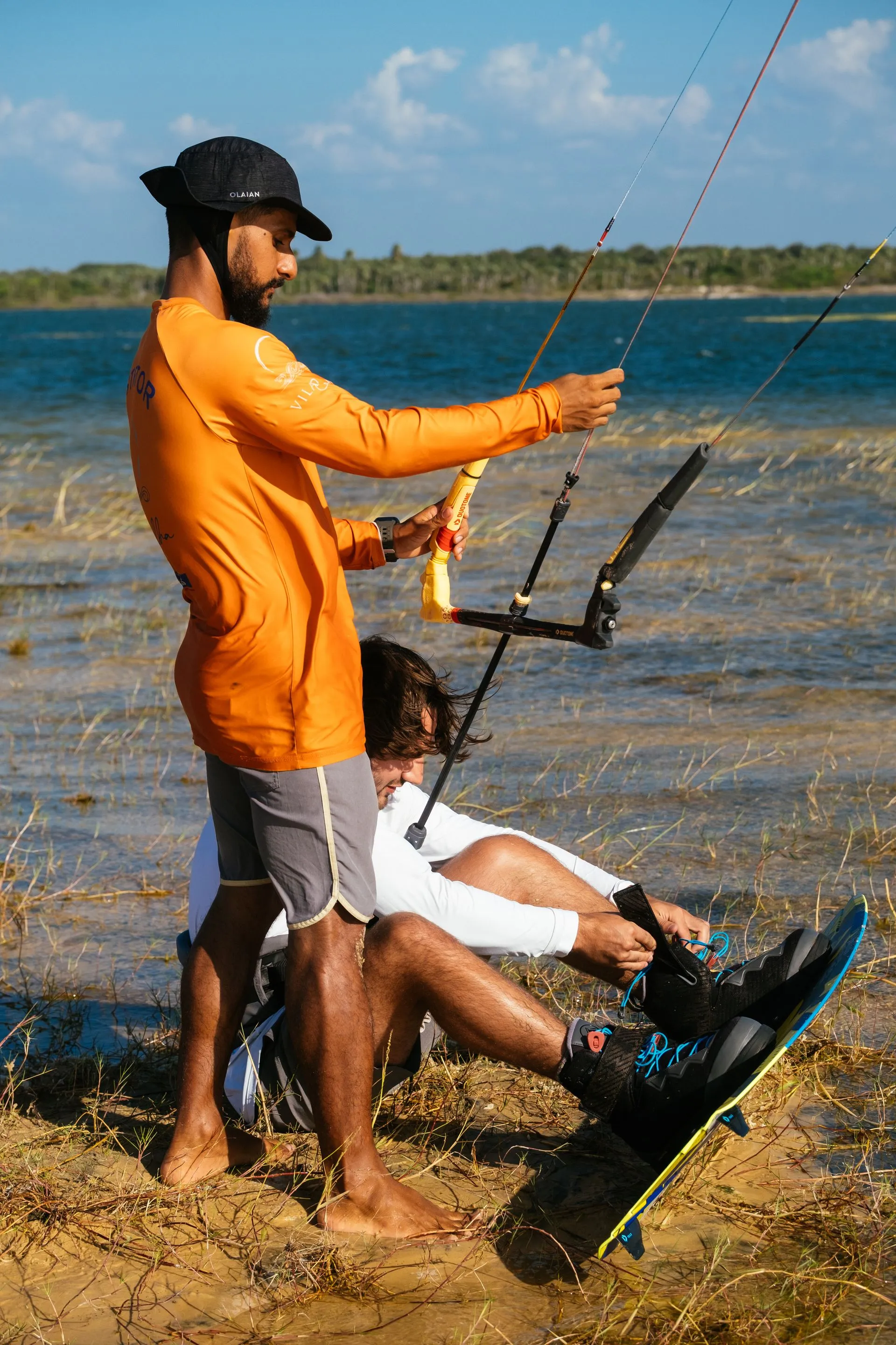 Instrutor segura as linhas do kite enquanto o aluno calça as botas da prancha na margem