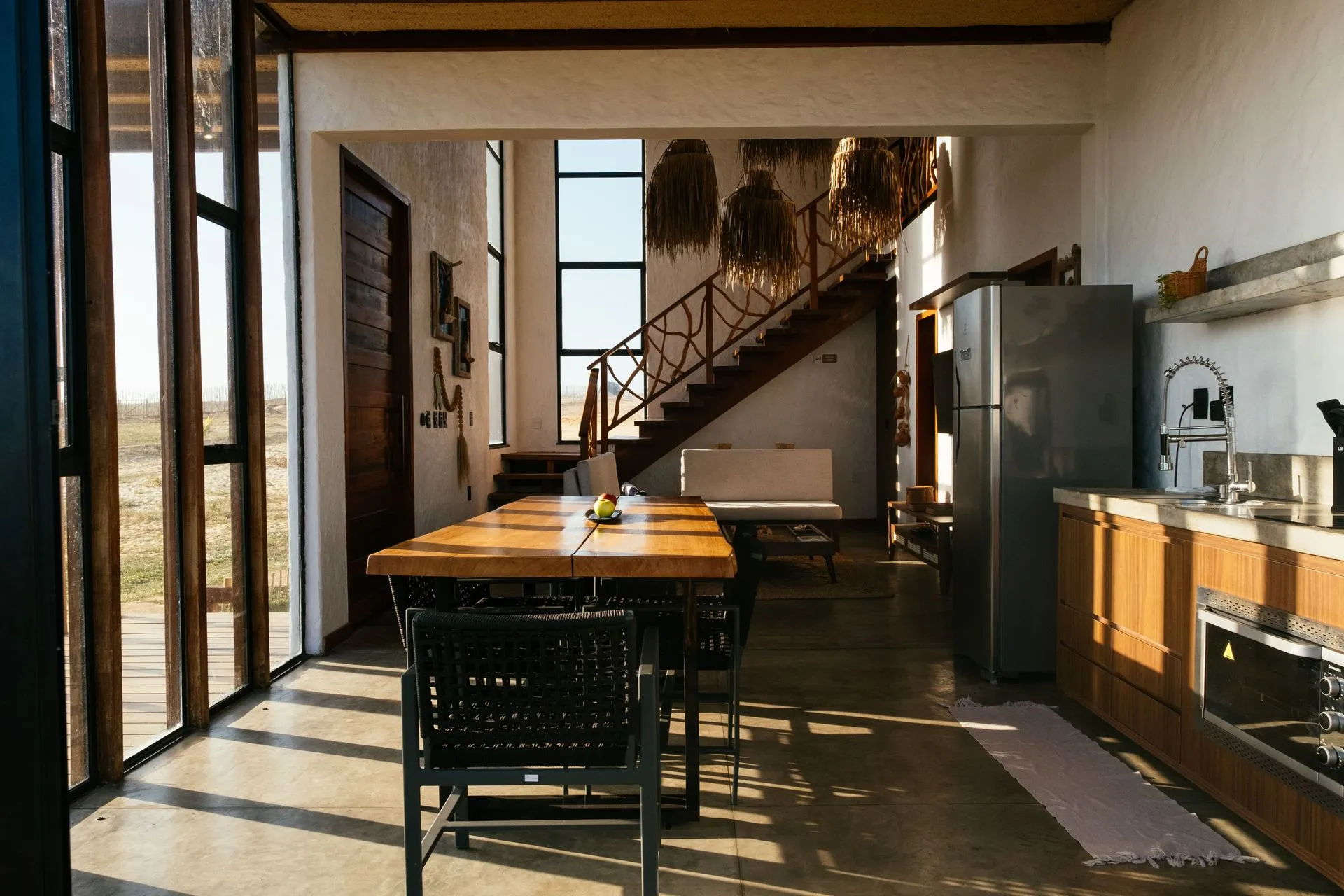 Cozinha e mesa de jantar integradas, com a escada de madeira ao fundo e luz da tarde nas janelas
