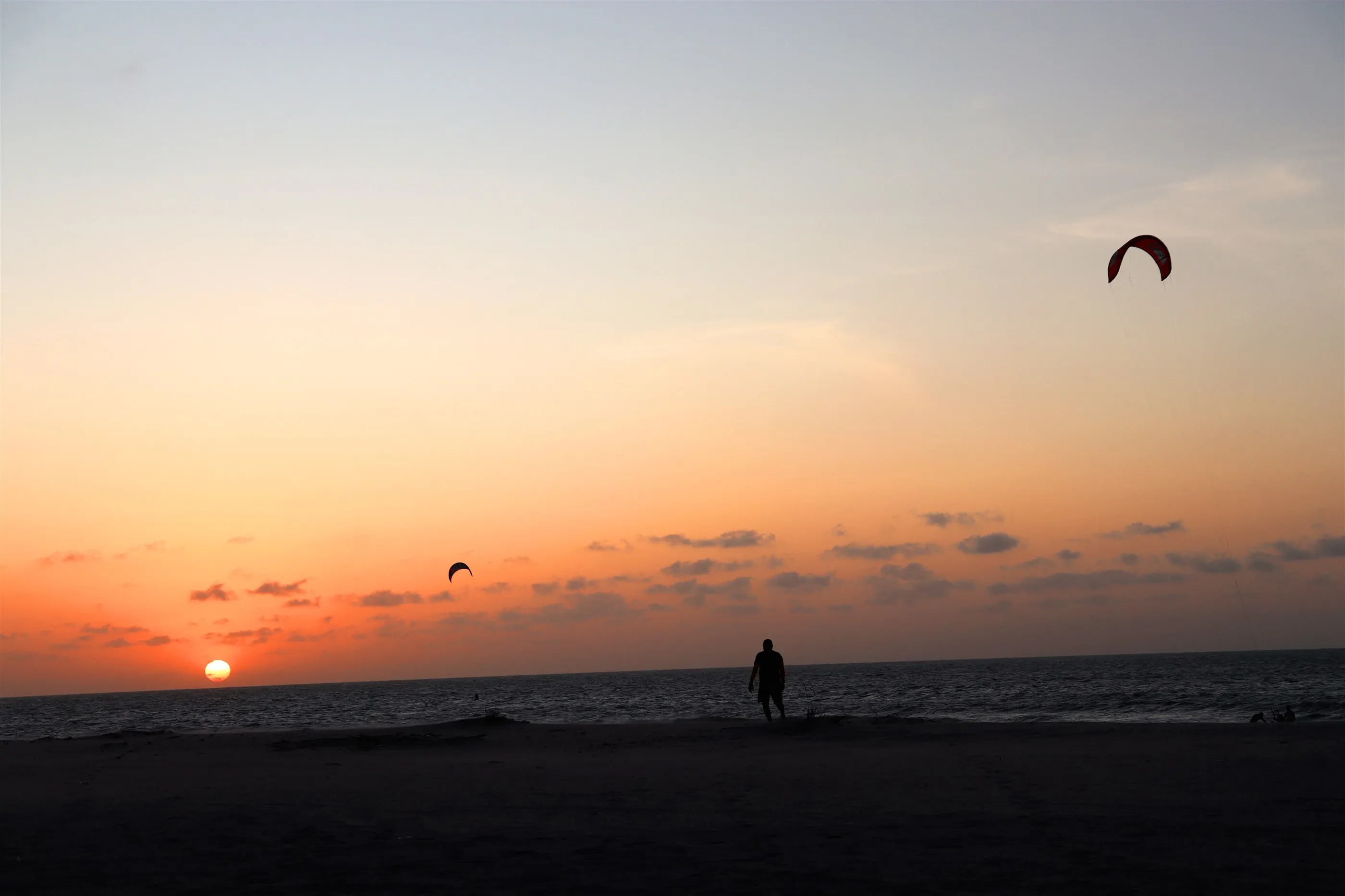 Silhueta de um kitesurfista na beira do mar ao pôr do sol, com kites espalhados pelo céu laranja