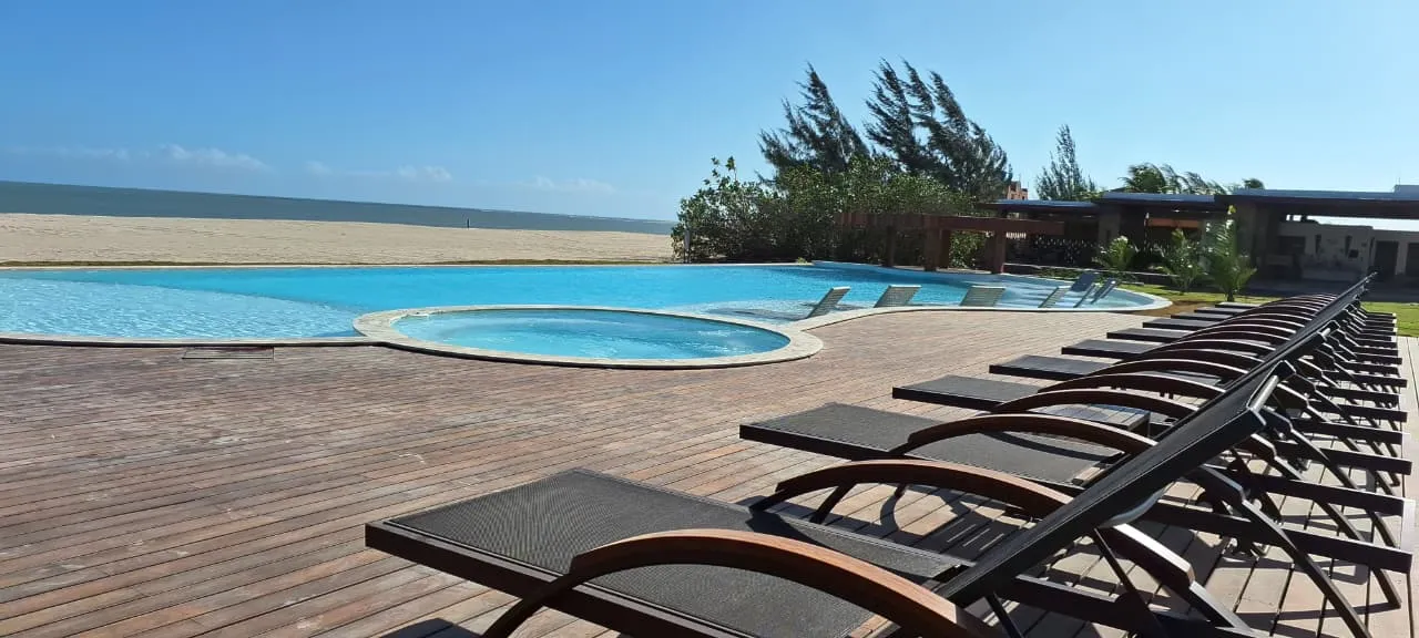 Fileira de espreguiçadeiras no deck de madeira, com a piscina, a hidromassagem redonda e a praia ao fundo