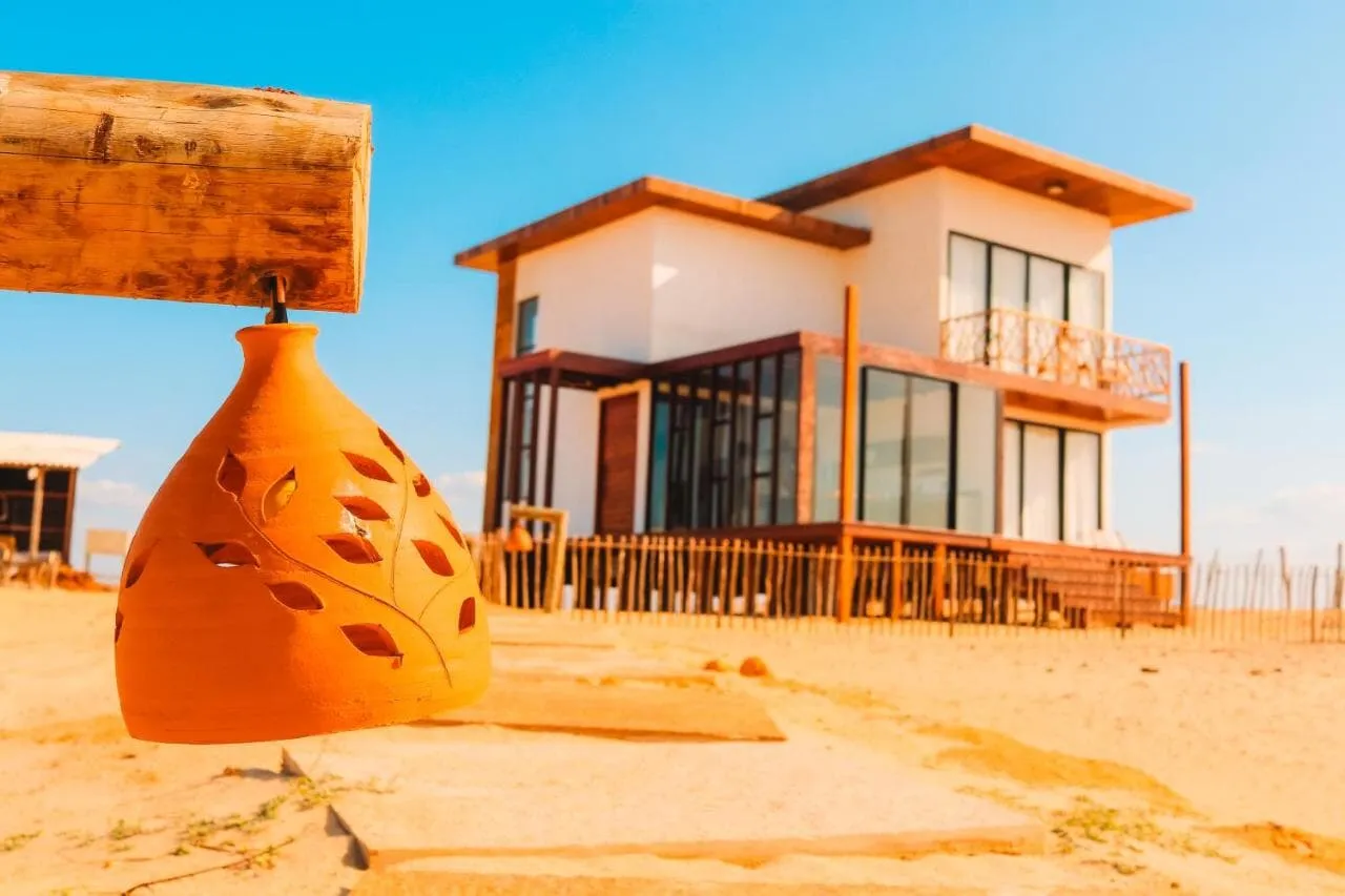 Fachada da casa de dois quartos erguida sobre a areia, com estrutura de madeira e cerca baixa