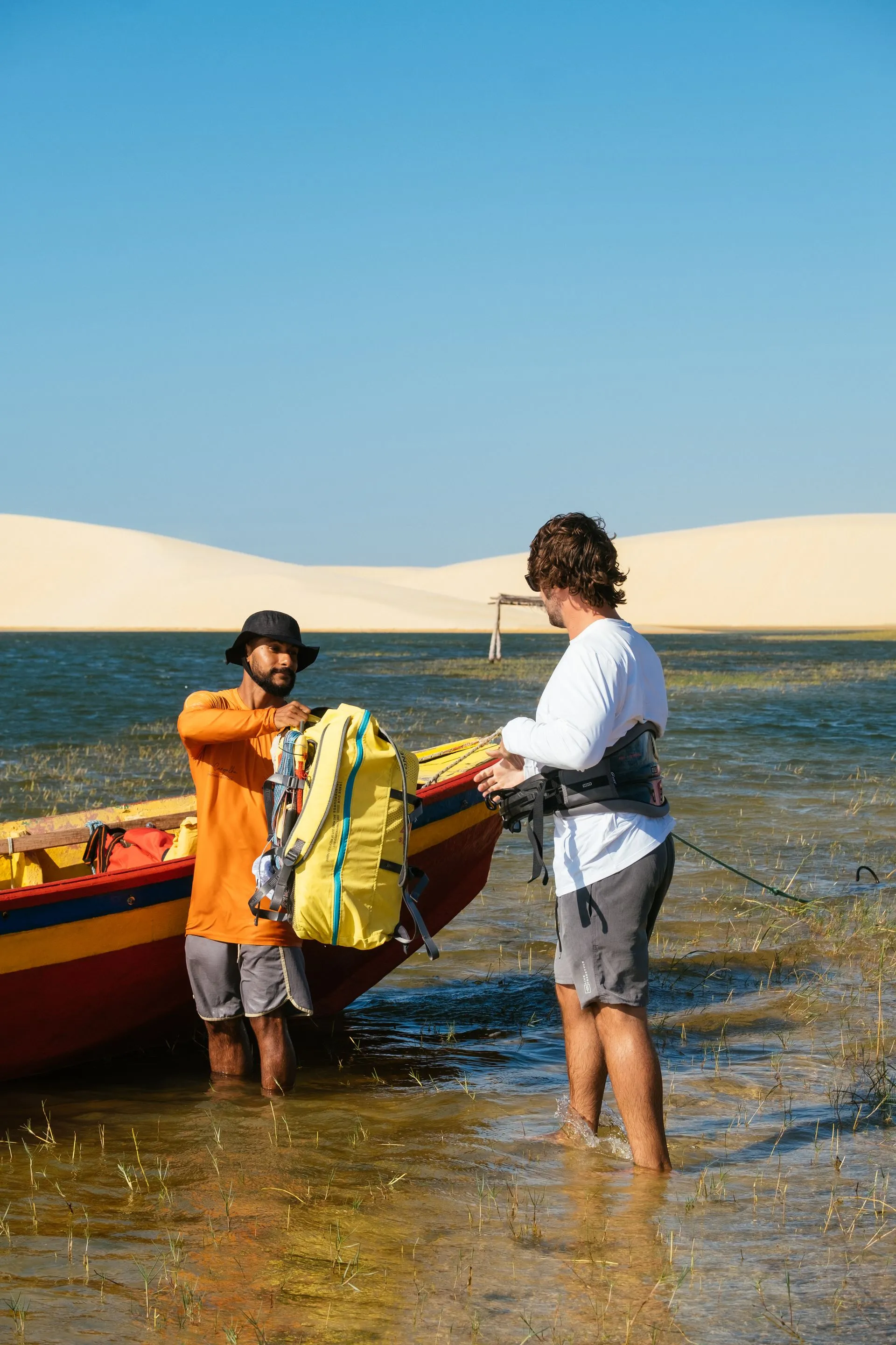 Instrutor entrega a mochila com o kite ao aluno ao lado de um barco colorido na lagoa