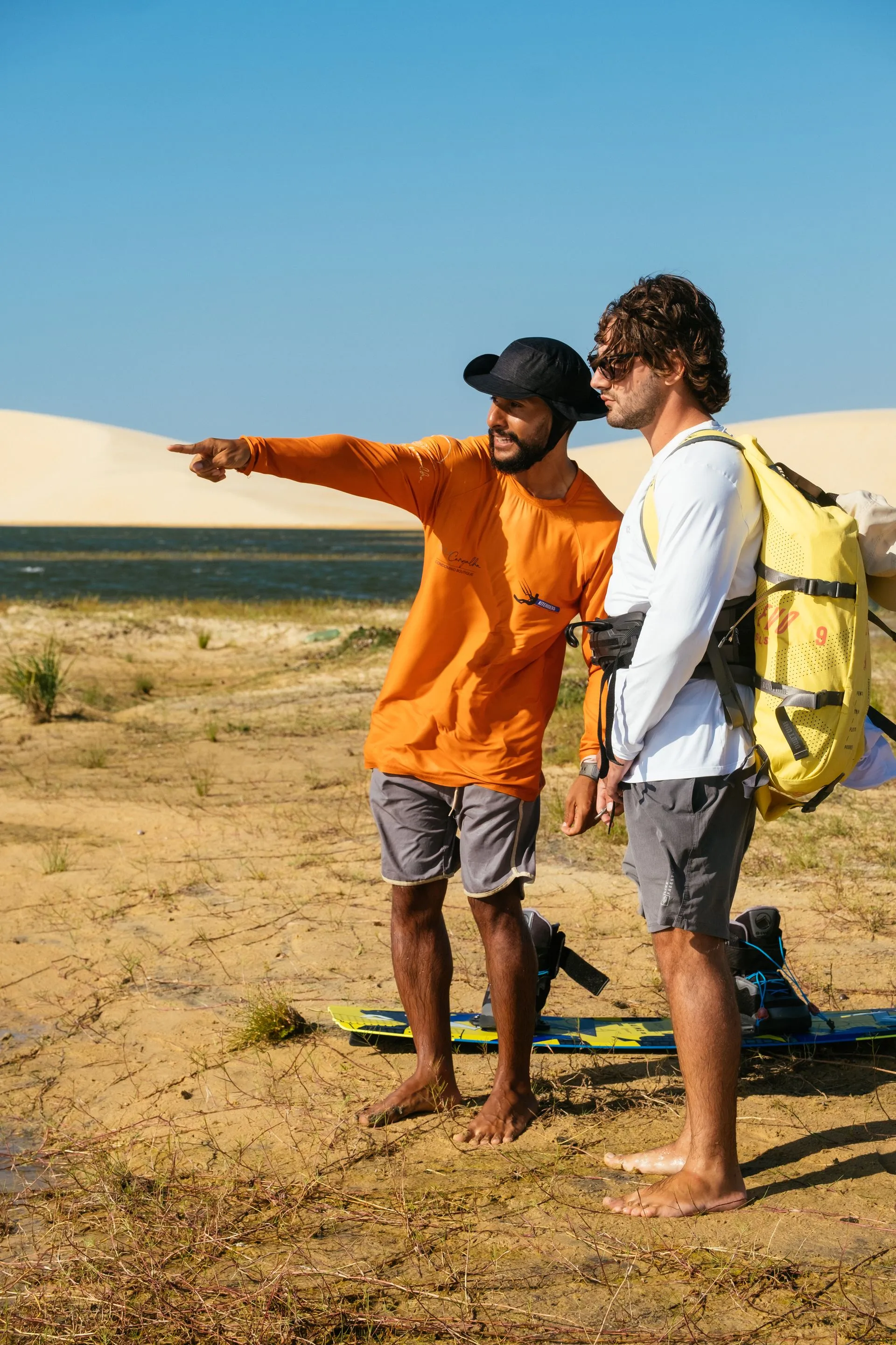 Instrutor aponta para a lagoa orientando o aluno na margem, com dunas ao fundo