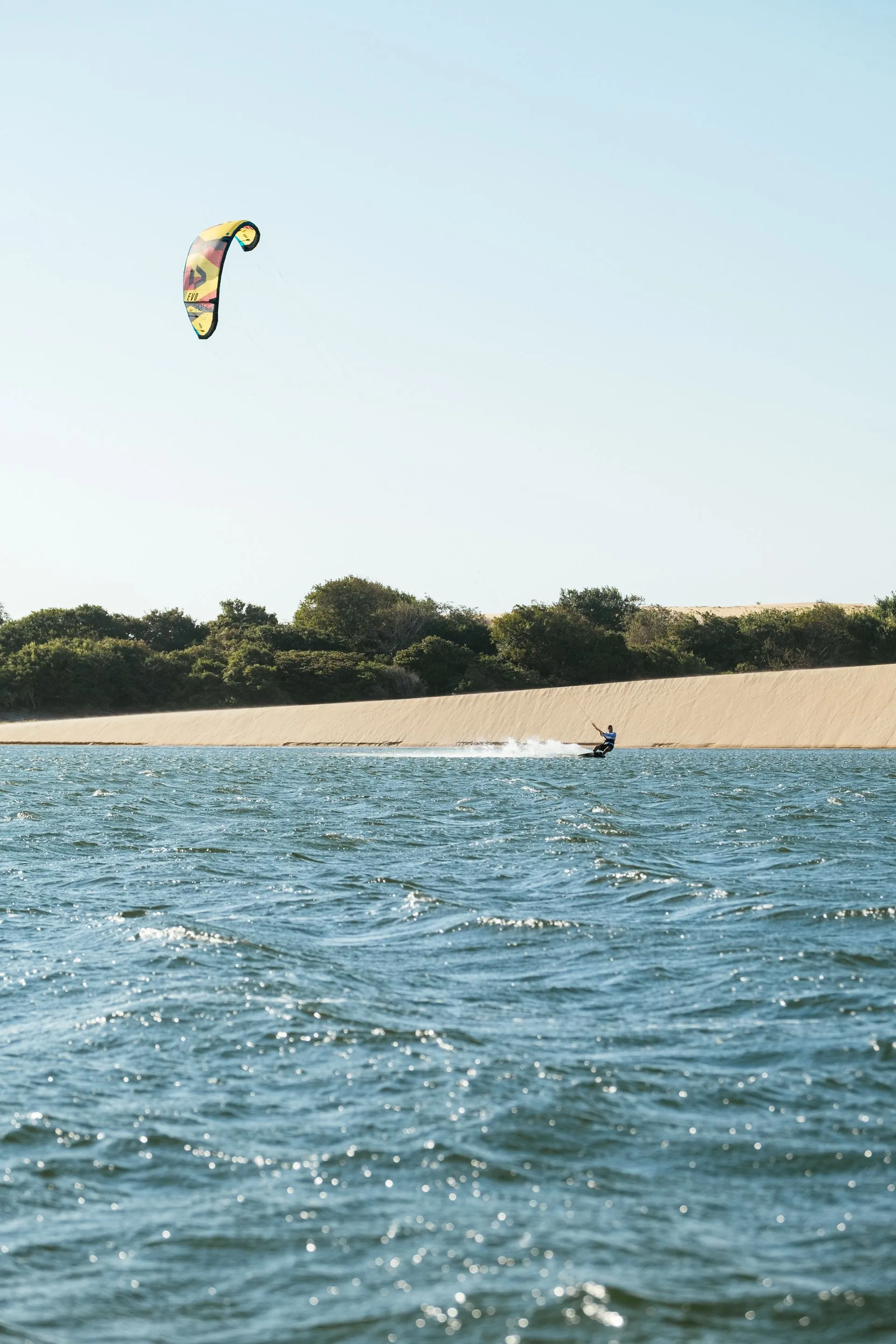 Kitesurfista com kite amarelo velejando em água agitada ao lado de uma duna e da vegetação