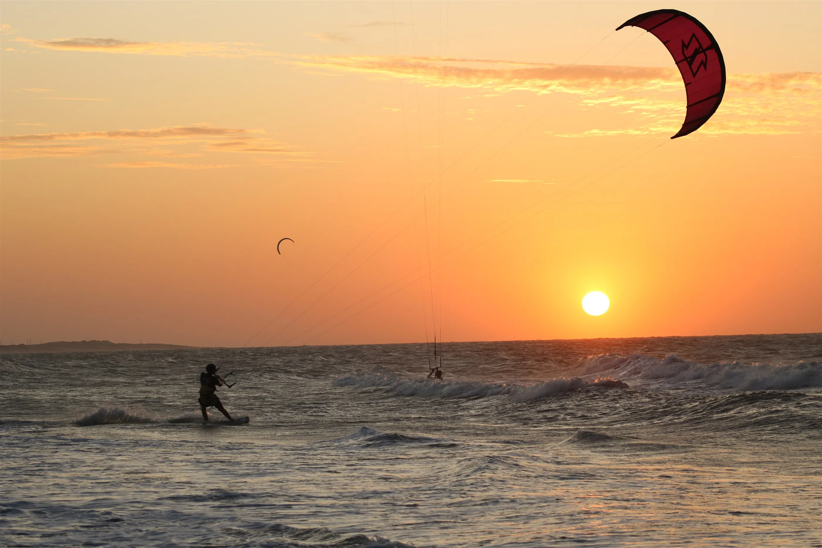 Kitesurfista velejando nas ondas ao pôr do sol, com um kite vermelho no alto do céu alaranjado