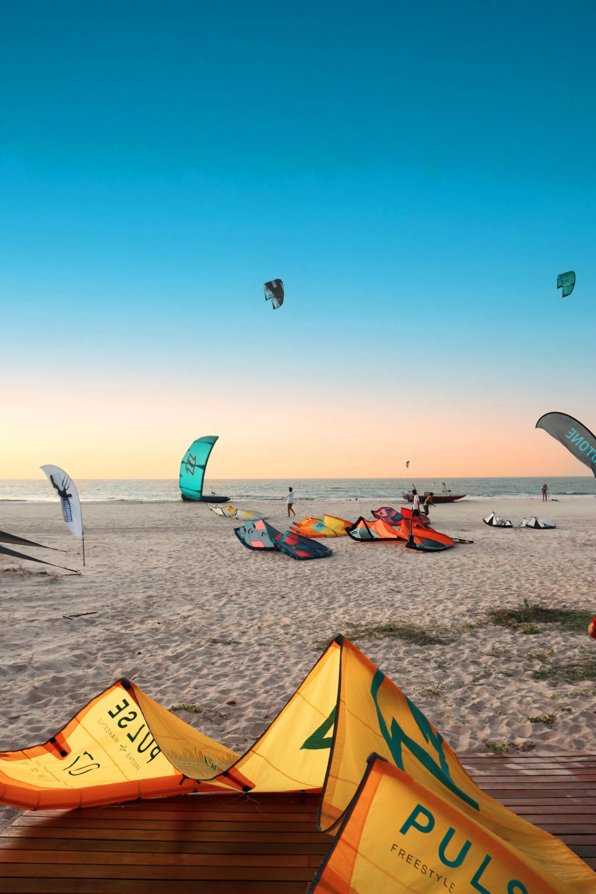 Kites coloridos espalhados na areia da praia ao entardecer, com outros kites no céu sobre o mar