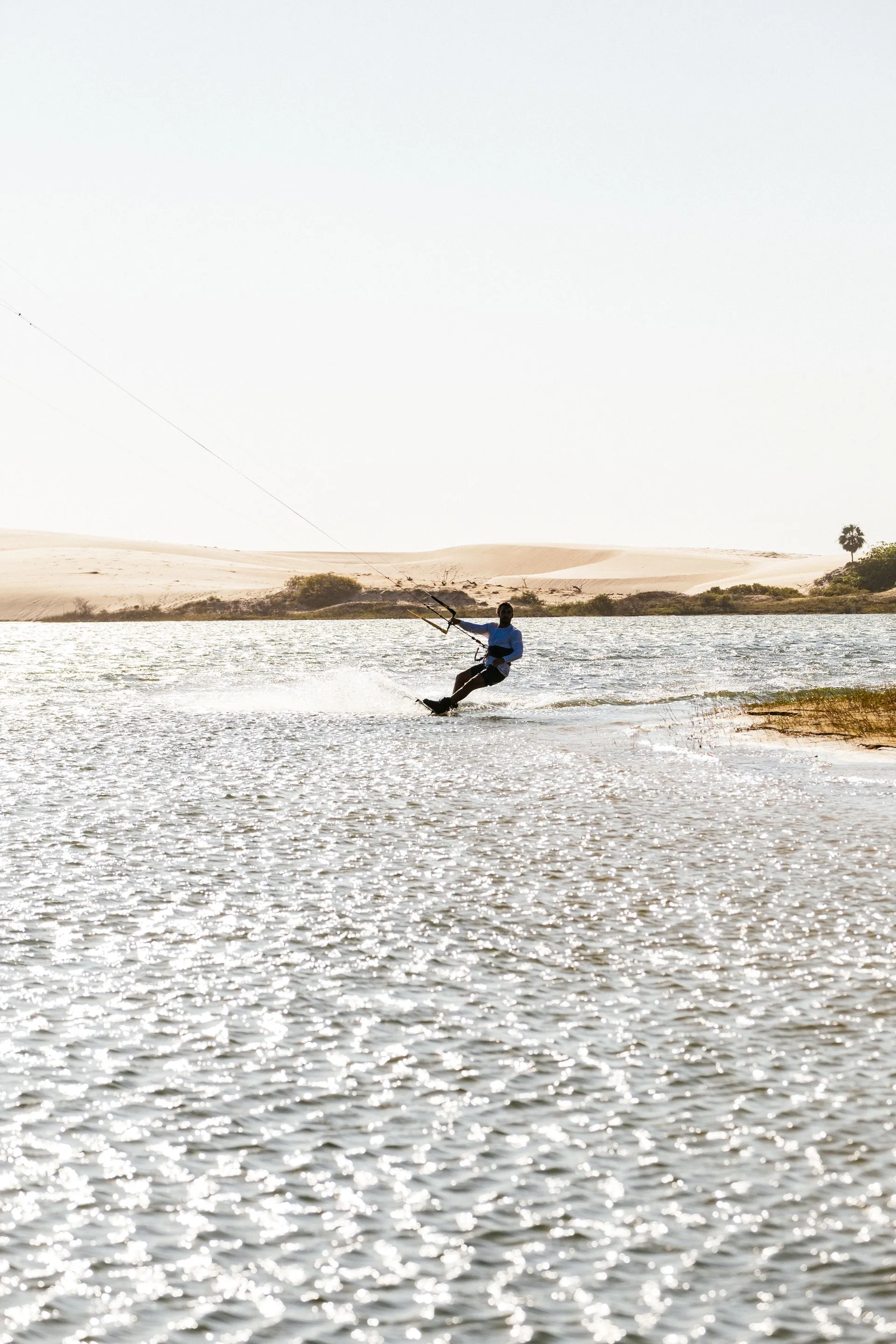 Kitesurfista deslizando em contraluz sobre a água brilhante da lagoa, com dunas ao fundo