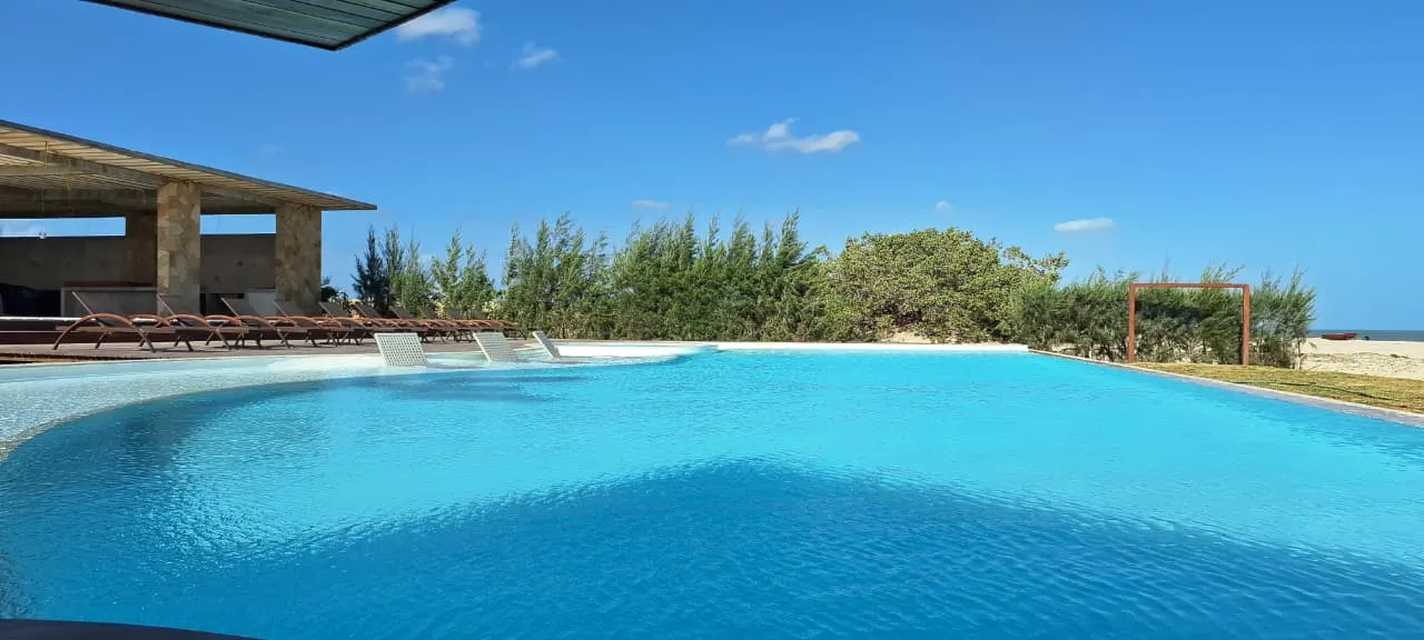 Piscina vista da beira da água, com espreguiçadeiras dentro da parte rasa e o terraço coberto à esquerda