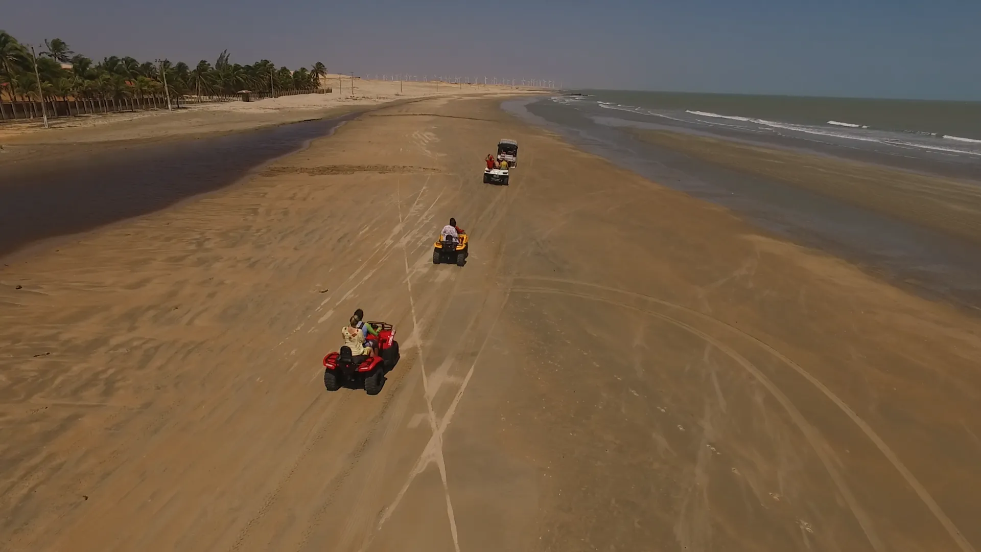 Quadriciclos percorrendo a faixa de areia entre o mar e os coqueirais, vistos do alto