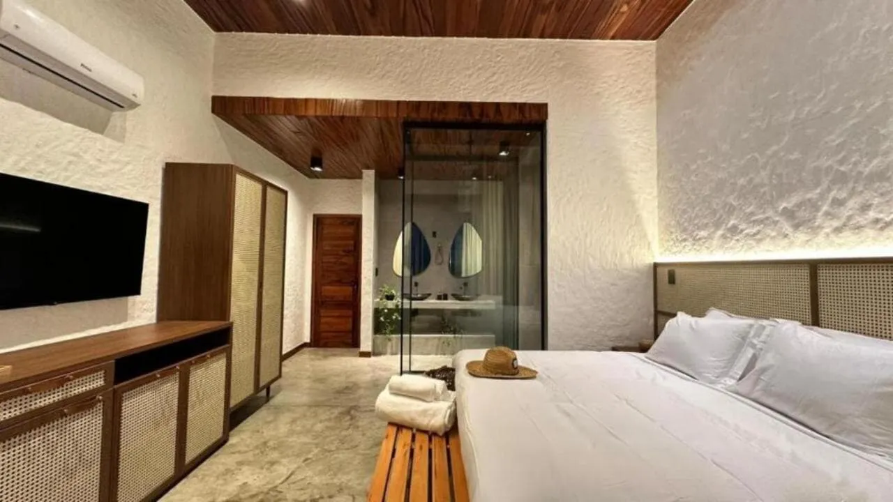 Quarto com televisão, armário de palhinha e banheiro de vidro ao fundo