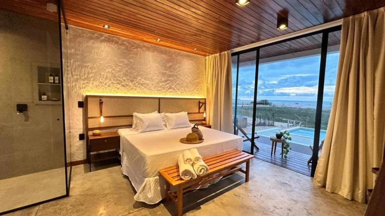 Quarto de casal com cabeceira iluminada e porta de vidro aberta para a varanda com vista para o mar